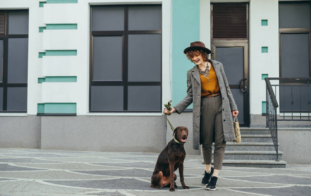 Smiling stylish woman and brown dog on a leash stand on the background of a building, girl smiling and looking at a pet.Fashionable portrait of a happy lady in a hat on a walk with puppies in the city - Photo, image
