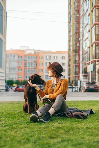 Stylish hipster girl sits on the lawn with a dog on a background of modern buildings and plays with a pet. Vertical portrait of a pet owner walking on a walk, sitting on a lawn and relaxing. - Photo, Image