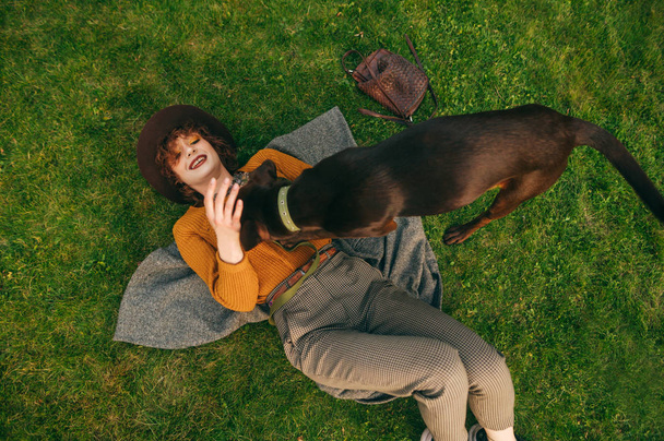 Усміхнена дівчина-хіпстер розважається з грайливою собакою, що лежить на зеленому газоні, вид зверху. Весела дівчина розслабляється на траві під час прогулянки з молодим собакою. Дозвілля з твариною
. - Фото, зображення