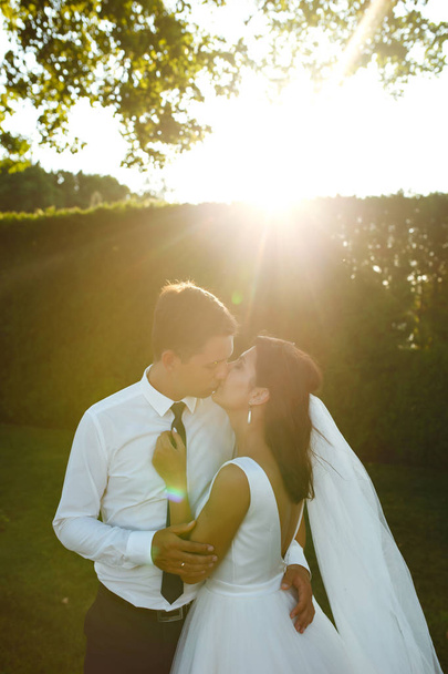 Lovely wedding couple at sunset. Bride and groom in wedding attire with a bouquet of flowers is in the hands against the backdrop of the green field at sunset. Romantic Married young family.  - Foto, immagini