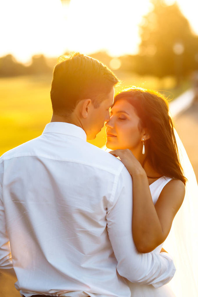 Υπέροχο ζευγάρι το ηλιοβασίλεμα. Νύφη και γαμπρός σε γαμήλια ενδυμασία με ένα μπουκέτο λουλούδια είναι στα χέρια με φόντο το πράσινο πεδίο στο ηλιοβασίλεμα. Ρομαντικό παντρεμένος νεαρή οικογένεια.  - Φωτογραφία, εικόνα