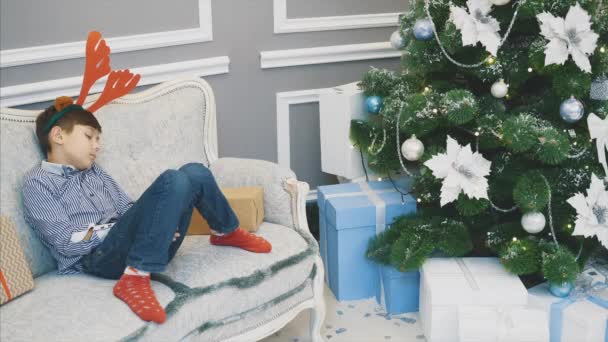 Lovely video of little boy with reindeer headgear, sleeping on the sofa among Christmas presenst near fir-tree. - Кадры, видео