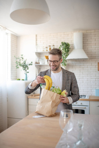 Man smiling while taking fruits and veggies out of bag - 写真・画像