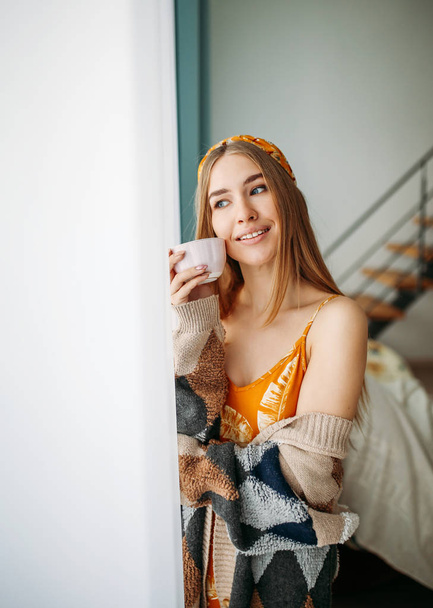 Όμορφη χαμογελαστή νεαρή γυναίκα δίκαιη μακριά μαλλιά κορίτσι φορώντας ένα άνετο πλεκτό ζακέτα με φλιτζάνι τσάι κοντά στο παράθυρο στο σπίτι - Φωτογραφία, εικόνα