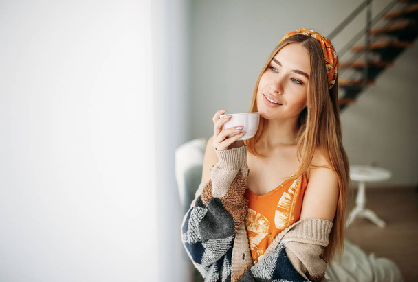 Piękny uśmiech młoda kobieta piękne długie włosy dziewczyna noszenie w przytulny sweter z filiżanką herbaty w pobliżu okna w domu - Zdjęcie, obraz