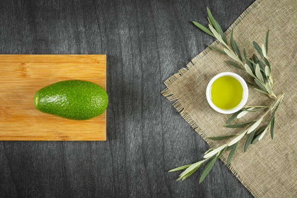 Healthy food concept. Fresh organic avocado, virgin olive oil and olive branches. Avocado with leafs on wooden table, top view, copy space. Natural ingredients. - Photo, Image