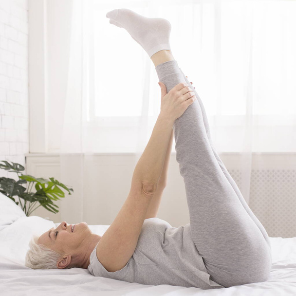 Senior woman doing morning gymnastics, stretching legs on bed - Photo, Image