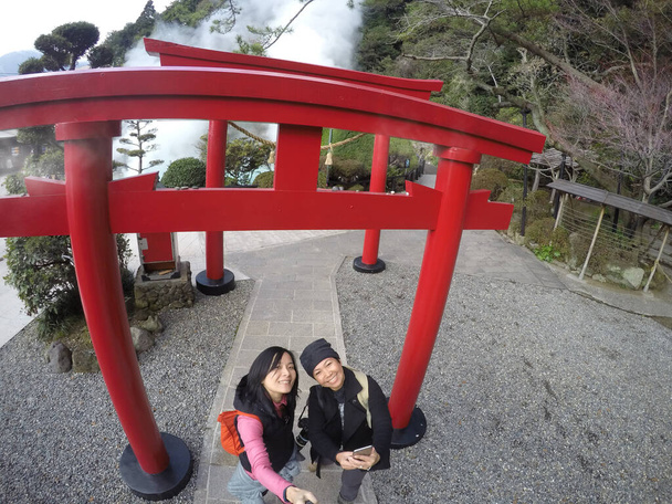 Two Asian tourist women take a photo under red wooden red shrine at Hot spring (Hell) onsen, hot blue water The "sea hell" in Umi Jigoku at Beppu, Oita-shi, Kyushu, Japan. Shooting with superwide angle, action camera. - Photo, Image