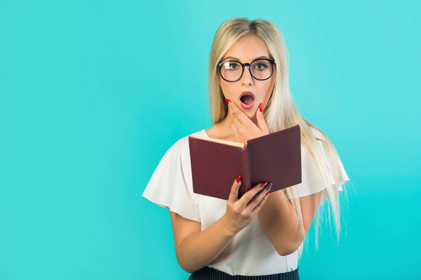 krásná mladá žena v brýlích drží knihu na modrém pozadí s překvapenou tváří - Fotografie, Obrázek