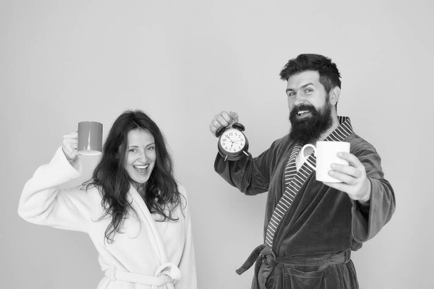 Every morning begins with coffee. Couple in bathrobes with mugs. Man with beard and sleepy woman enjoy morning coffee or tea. Time to wake up and have nice day. Guy husband hold coffee and clock - Foto, Bild