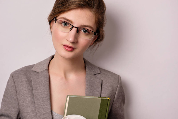 A close portrait of a girl in glasses and a gray jacket with books in her hands looking into the frame, on a light background. - Фото, изображение