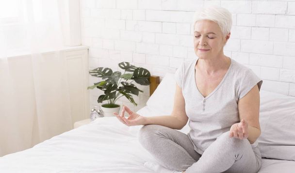 Femme âgée faisant du yoga, méditant en position lotus sur le lit à la maison
 - Photo, image
