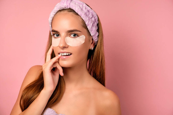 A girl in a purple towel stands on a pink background with patches under her eyes and looks into the frame with her bite of finger. - Foto, imagen