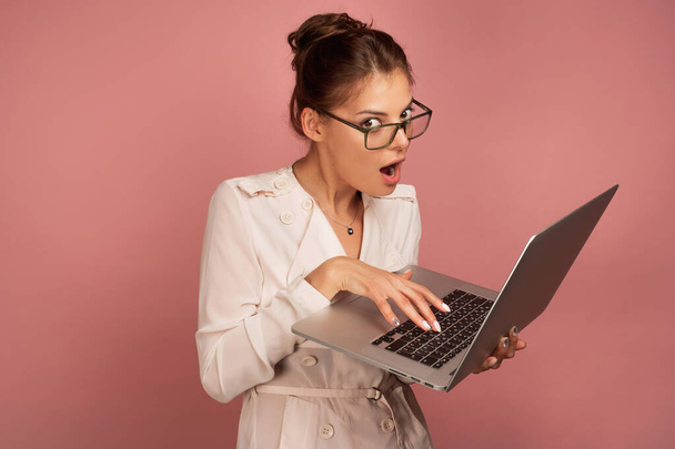 The girl in glasses in a light jacket on a pink background stands typing on a laptop with a funny face. - Foto, imagen