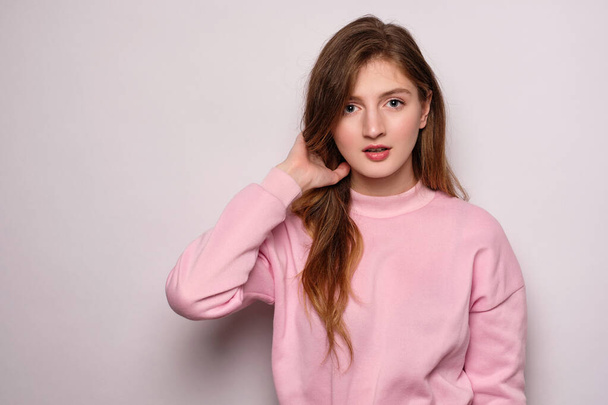 A blonde girl in a pink sweatshirt stands on a white background straightening her hair looks into the frame - Fotoğraf, Görsel
