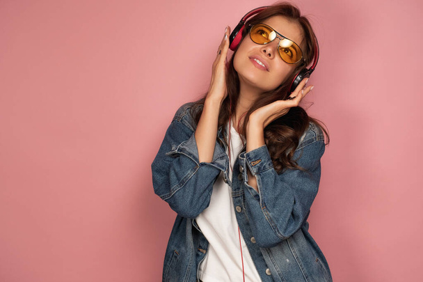 A brunette on a pink background in jeans looks dreamily up and holds her headphones. - Фото, зображення
