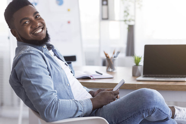 Retrato de un alegre empleado afroamericano usando un teléfono inteligente en el lugar de trabajo
 - Foto, imagen