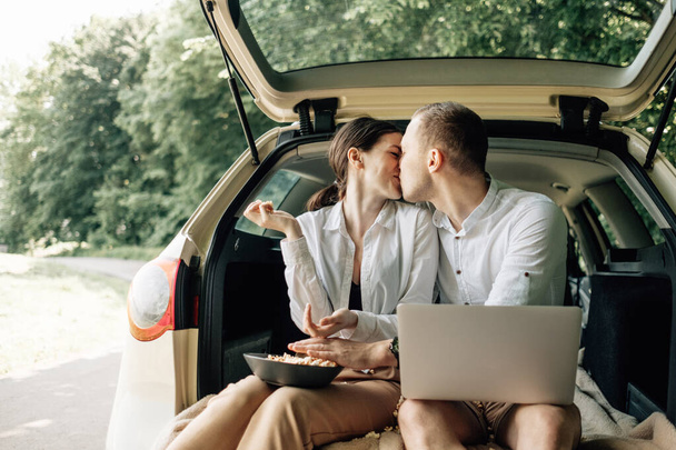 junges glückliches Paar in weißem T-Shirt im Kofferraum sitzend mit Laptop und Popcorn am Straßenrand, Wochenende außerhalb der Stadt, Urlaub und Roadtrip-Konzept - Foto, Bild