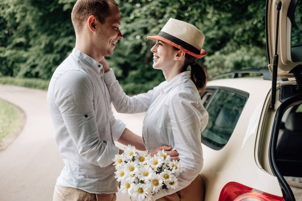 Jovem casal feliz vestido como em T-shirt branca se divertindo perto do carro, fim de semana fora da cidade, feriados e conceito de viagem rodoviária
 - Foto, Imagem