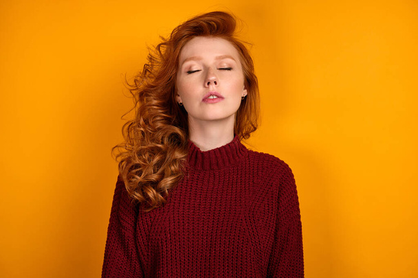 A red-haired girl with a shock of curls in a burgundy sweater stands on a yellow background with her eyes closed. - Foto, immagini
