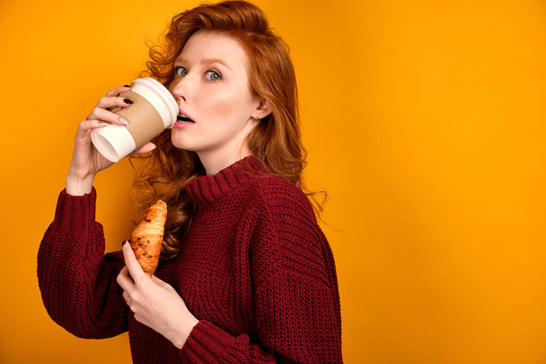 Red-haired curly girl in a sweater stands half-turn takes a sip from a paper cup while holding a croissant, looks into the frame - Foto, afbeelding