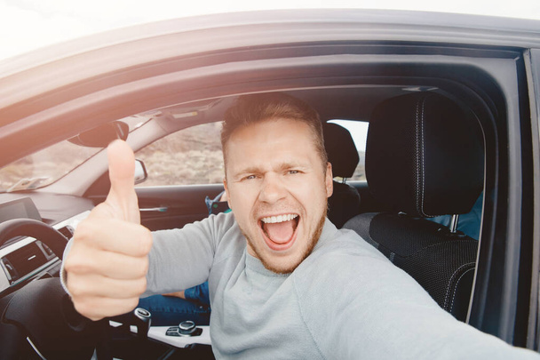Driver of car takes selfie photo, shows thumb up from window. Man smiles, mouth wide open, sunlight. Travel rental concept - Foto, afbeelding