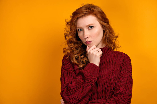 Red-haired curly girl in a burgundy sweater on yellow background looks thoughtfully into the frame holding her chin with her hand - Photo, Image