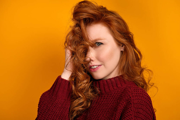 Beautiful red-haired girl in a sweater looks at the camera smiling while straightening a pile of hair on a yellow background - Photo, Image