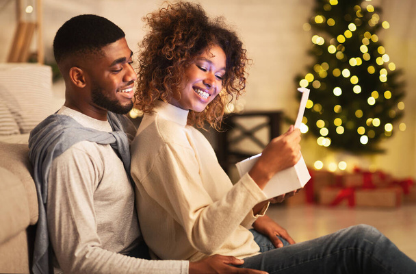 Ενθουσιασμένο αφροαμερικανό ζευγάρι που ανοίγει χριστουγεννιάτικο δώρο στο σαλόνι, μπροστά από το χριστουγεννιάτικο δέντρο - Φωτογραφία, εικόνα
