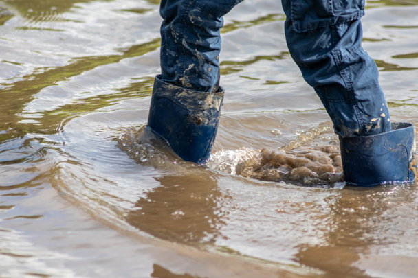 Jonge jongen met korte blauwe trowsers waden met natte sokken en natte laarzen bij hoogwater nadat een overstromingswater de dijk heeft doorbroken en de landen achter zich heeft overgevlogen - Foto, afbeelding