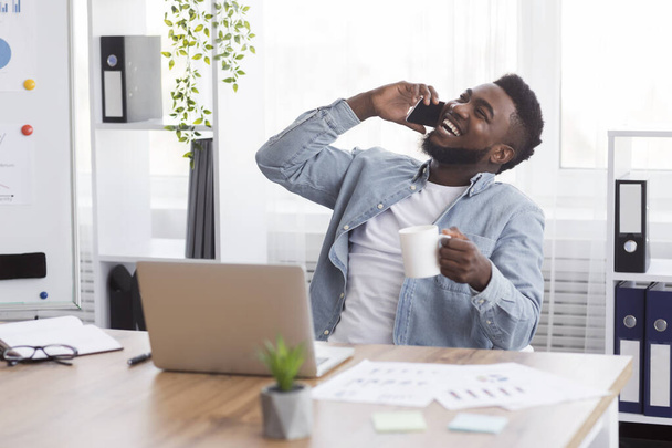 Radosny afrykański biznesmen rozmawiający przez telefon i pijący kawę w miejscu pracy - Zdjęcie, obraz