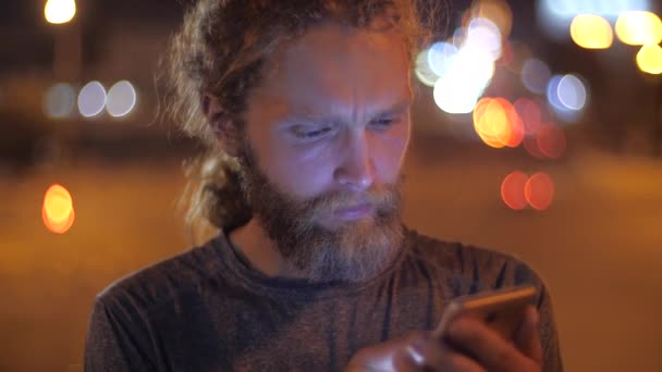 Close-up, portret van een knappe blanke bebaarde langharige jonge man die smartphone gebruikt op de achtergrond van het verplaatsen van Avondverlichting van een stad. - Video