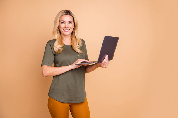 Фото веселой позитивной милой довольно милой, зубастой зеленой сияющей девушки, держащей ноутбук с руками в желтых брюках, просматривающих изолированный пастельно-бежевый цвет фона
 - Фото, изображение