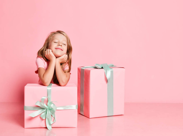 Christmas and New Year. Kid girl is sitting behind gifts with her chin in her hands, dreaming with closed eyes on pink - Foto, afbeelding