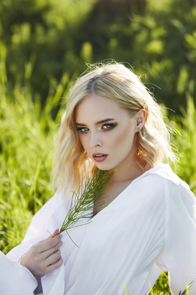 Girl in a long white dress sits on the grass in a field. Blonde  - Foto, afbeelding