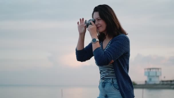 young woman takes pictures on a vintage camera by the ocean. portrait of a girl with a retro camera - Πλάνα, βίντεο