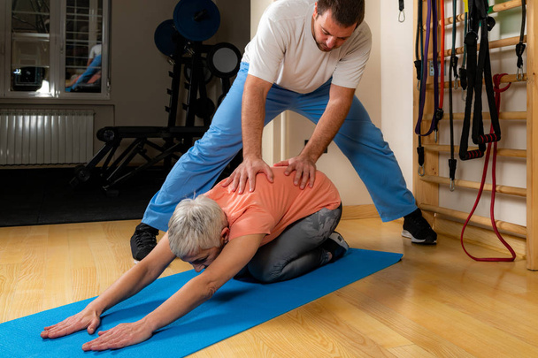 Fizjoterapeuta lub trener rozciągający starszą kobietę na macie do ćwiczeń w siłowni. - Zdjęcie, obraz