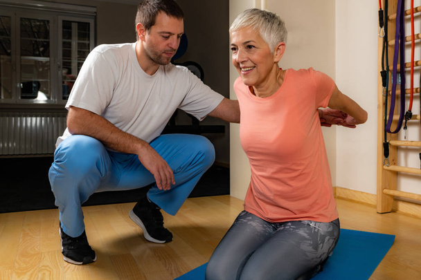 Фізіотерапевт або тренер, що розтягує старшу жінку на тренувальному килимку в спортзалі
. - Фото, зображення