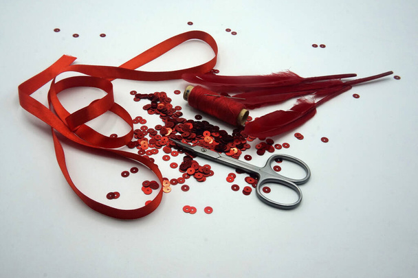 rote Pailletten, Spule aus rotem Faden, Nadel, Schere, rotes Band und rote Federn auf weißem Hintergrund. - Foto, Bild