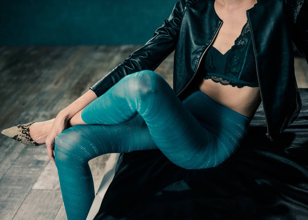 young stylish woman in colorful leggins and leather jacket sitting on black bed sheet - 写真・画像