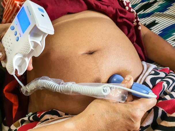 ακούγοντας τον ήχο της καρδιάς του εμβρύου σε μια έγκυο γυναίκα κοιλιά από ένα εμβρυϊκό μηχάνημα doppler από μια εκπαιδευμένη γυναίκα εργαζόμενος υγείας στο ινδικό σπίτι - Φωτογραφία, εικόνα