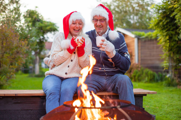 Senior couple with Christmas santa's hats and hot drinks in front of a fireplace or fireside - Foto, afbeelding
