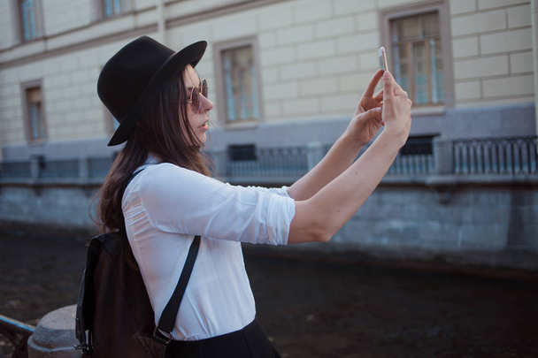 Tourist in the city takes a photo on smartphone. Young woman in black hat and white shirt. - Fotografie, Obrázek