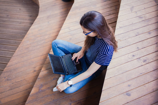 A brunette girl, a student with long hair, with glasses sitting cross-legged on wooden steps and typing on laptop. - Foto, afbeelding