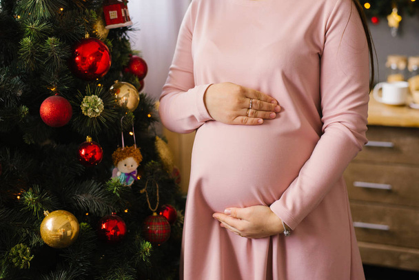 Frau hält Hände auf ihrem schwangeren Bauch vor dem Hintergrund der Weihnachtsbaumbeleuchtung. - Foto, Bild