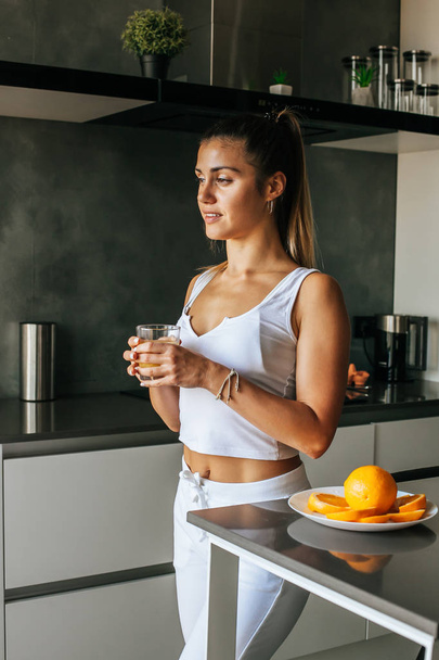 Young athletic girl drinks fresh orange juice in the morning in the kitche - Zdjęcie, obraz