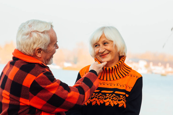 Vrolijke ouderen een vrouw en een man zitten op de stenen en knuffelen op het meer, tegen de achtergrond van de brug. - Foto, afbeelding