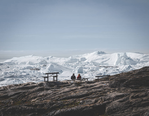 Two people sitting with view towards Icefjord in Ilulissat. Easy hiking route to the famous Kangia glacier in Greenland. The Ilulissat Icefjord seen from the viewpoint. - Foto, afbeelding