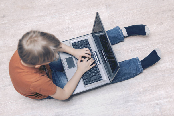 ein Laptop auf den Füßen eines Jungen, der auf dem Boden sitzt. Die Hände sind auf der Tastatur. Computerspiele spielen, trainieren, bloggen, tippen. Ansicht von oben. Selektiver Fokus - Foto, Bild