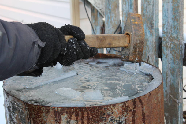 un hombre rompe el hielo con un martillo en un barril / foto agua congelada en un tanque barrel.the es metal.the agua estaba helada cold d.a hombre rompe el hielo en la superficie del water.works con un martillo
. - Foto, Imagen